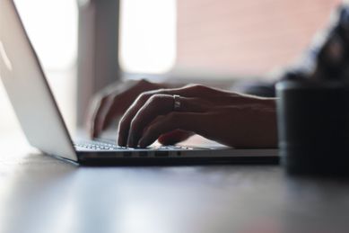 En person med en ring på venstre hånd som sitter ved et bord og skriver på en laptop med lys som kommer inn fra vinduet i bakgrunnen