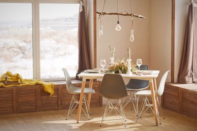 Interiør i en lys og varm spisestue som består av et dekket på hvitt spisebord med tre bein, 4 stoler og store vindu som slipper inn mye naturlig lys, moderne møbler