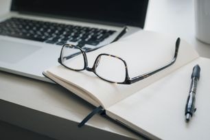 Briller med svart ramme som ligger på en notatbok med en penn ved siden av en laptop på et skrivebord i et lyst rom